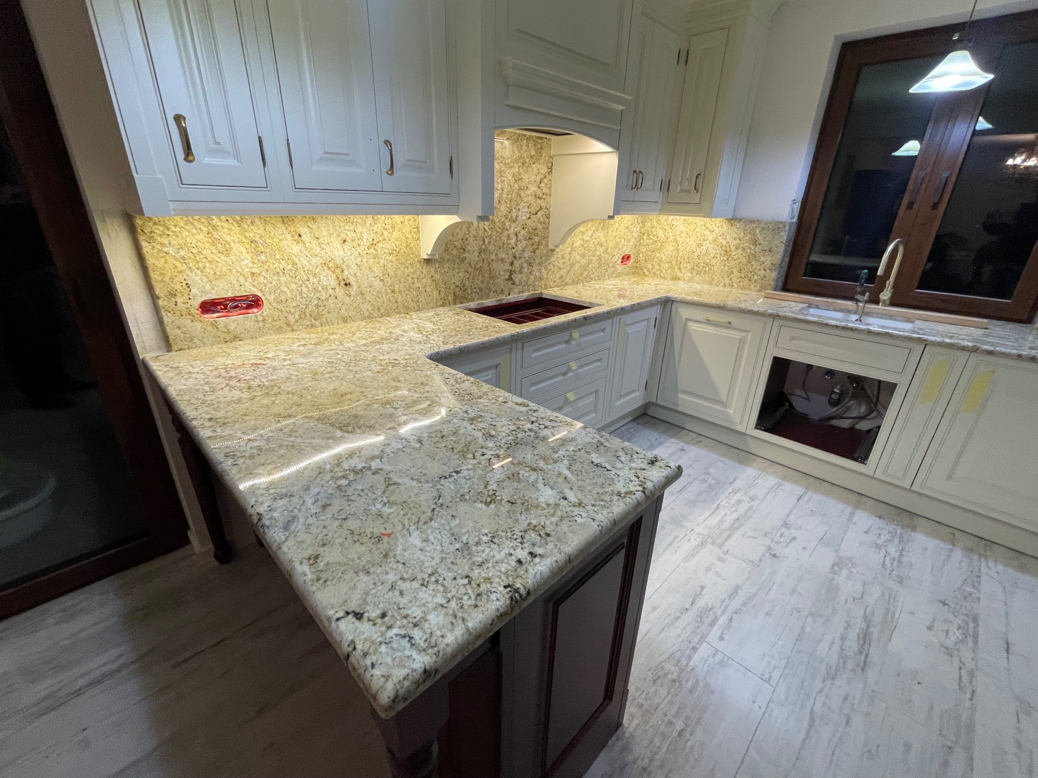 Jasny blat kamienny z wyspą w klasycznej kuchni z białymi szafkami i oknem. Widoczne detale wykończenia i oświetlenie podszafkowe.