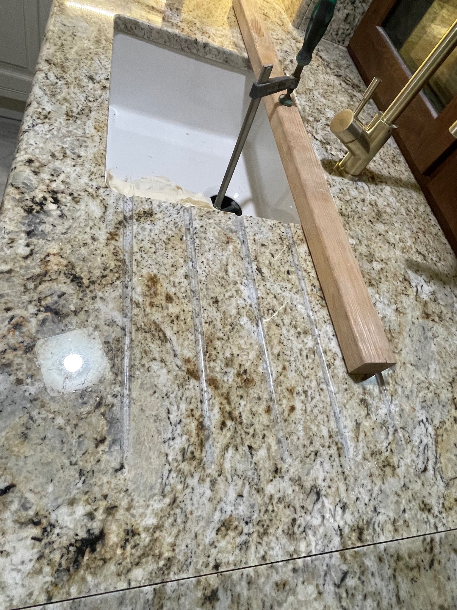 Montaż blatu kamiennego: zlew w wyciętym otworze, zabezpieczony drewnianą listwą i ściskiem. Widoczna bateria i fragment szafki. Blat z jasnego kamienia z ciemnymi plamkami.