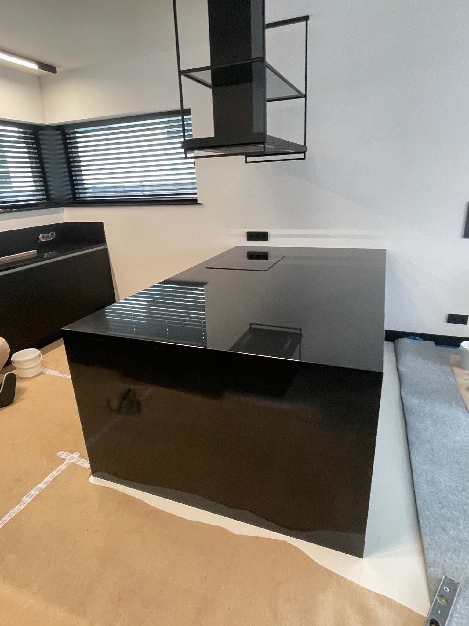 Czarny, błyszczący blat kuchenny z wbudowaną płytą grzewczą, widoczny w nowoczesnej kuchni z minimalistycznym okapem i żaluzjami w oknie.