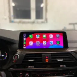 Wnętrze samochodu BMW z widocznym ekranem dotykowym prezentującym interfejs CarPlay z ikonami aplikacji, w tle widoczne okno.