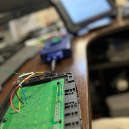 Codena Motors Patryk Płomiński - Zielona płytka drukowana z podłączonymi kolorowymi przewodami, podłączona do niebieskiego urządzenia diagnostycznego, na drewnianym biurku z rozmytym tłem ekranu diagnostycznego.