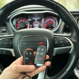 Codena Motors Patryk Płomiński - Dwie różne wersje kluczyków samochodowych Dodge trzymane w dłoni na tle kierownicy Dodge Challenger z widocznym logo i deską rozdzielczą.