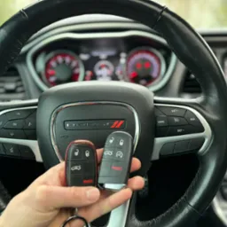 Dwie różne wersje kluczyków samochodowych Dodge trzymane w dłoni na tle kierownicy Dodge Challenger z widocznym logo i deską rozdzielczą.