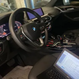 Wnętrze samochodu BMW z włączonym laptopem wyświetlającym diagnostykę pojazdu, widoczne deska rozdzielcza, kierownica z logo BMW i ekran nawigacji.