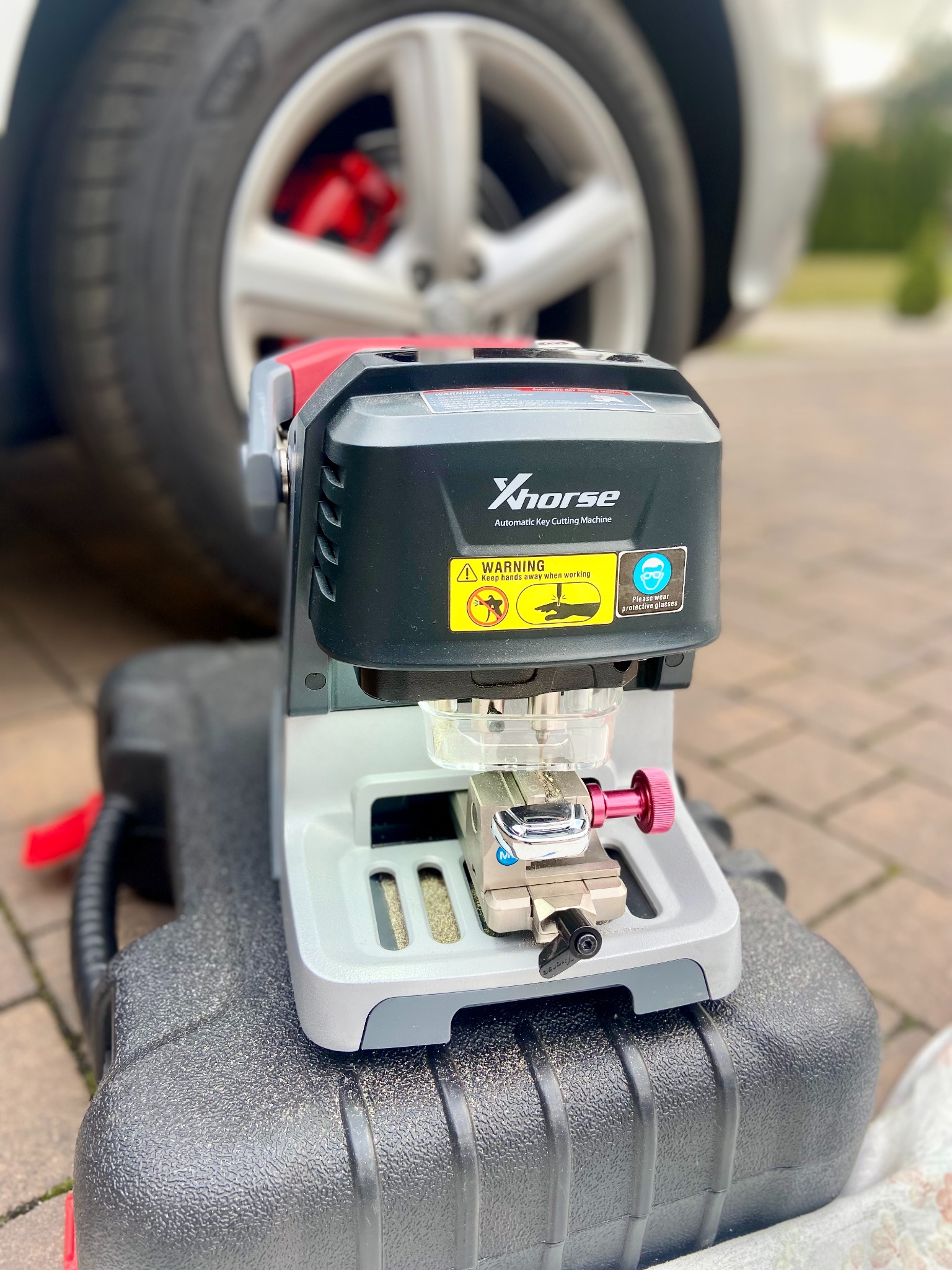 Automatyczna maszyna do cięcia kluczy samochodowych Xhorse na czarnym pudełku transportowym, w tle koło samochodu z czerwonym zaciskiem hamulcowym.