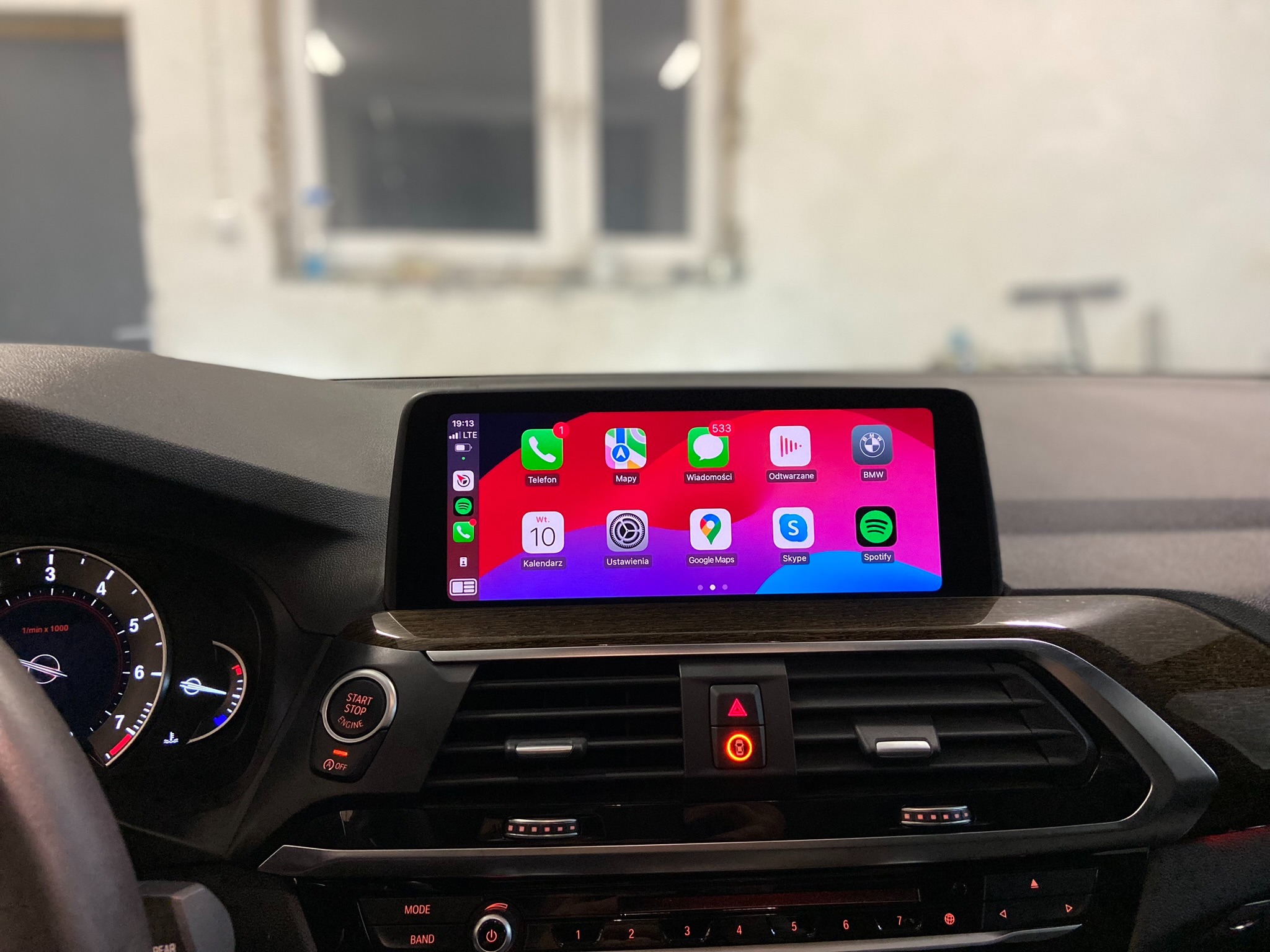 Wnętrze samochodu BMW z widocznym ekranem dotykowym prezentującym interfejs CarPlay z ikonami aplikacji, w tle widoczne okno.
