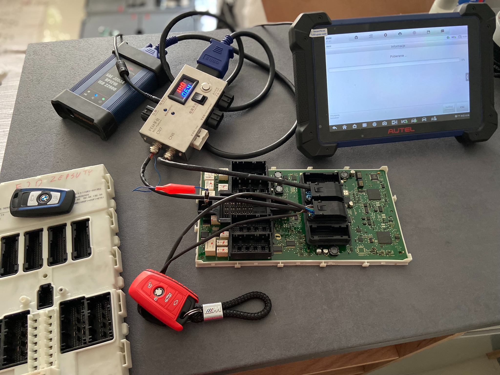 Diagnostyka sterownika samochodowego BMW za pomocą testera diagnostycznego Autel, z widoczną płytką drukowaną, przewodami i kluczykami, wskazując na proces pobierania informacji.