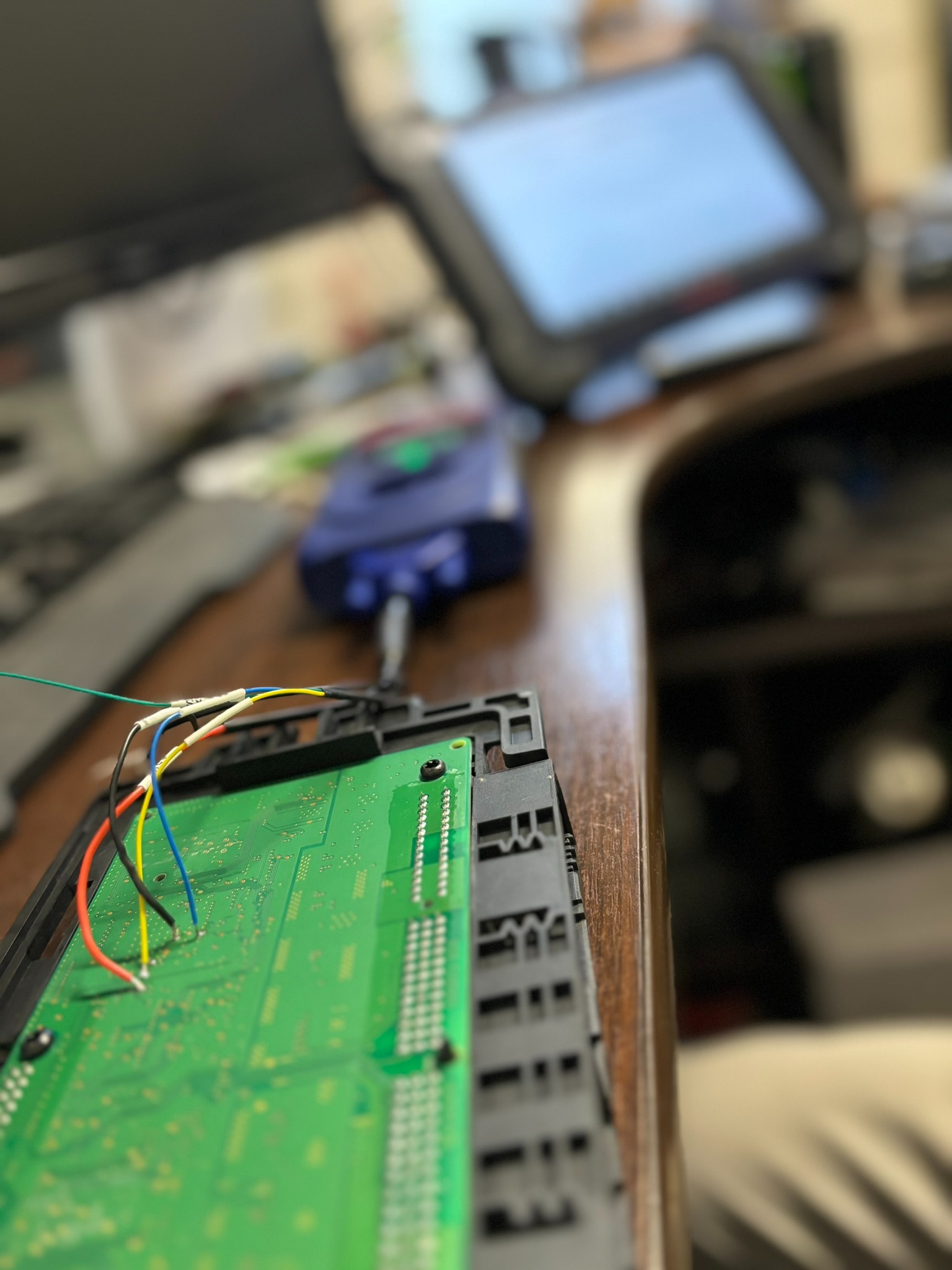 Zielona płytka drukowana z podłączonymi kolorowymi przewodami, podłączona do niebieskiego urządzenia diagnostycznego, na drewnianym biurku z rozmytym tłem ekranu diagnostycznego.