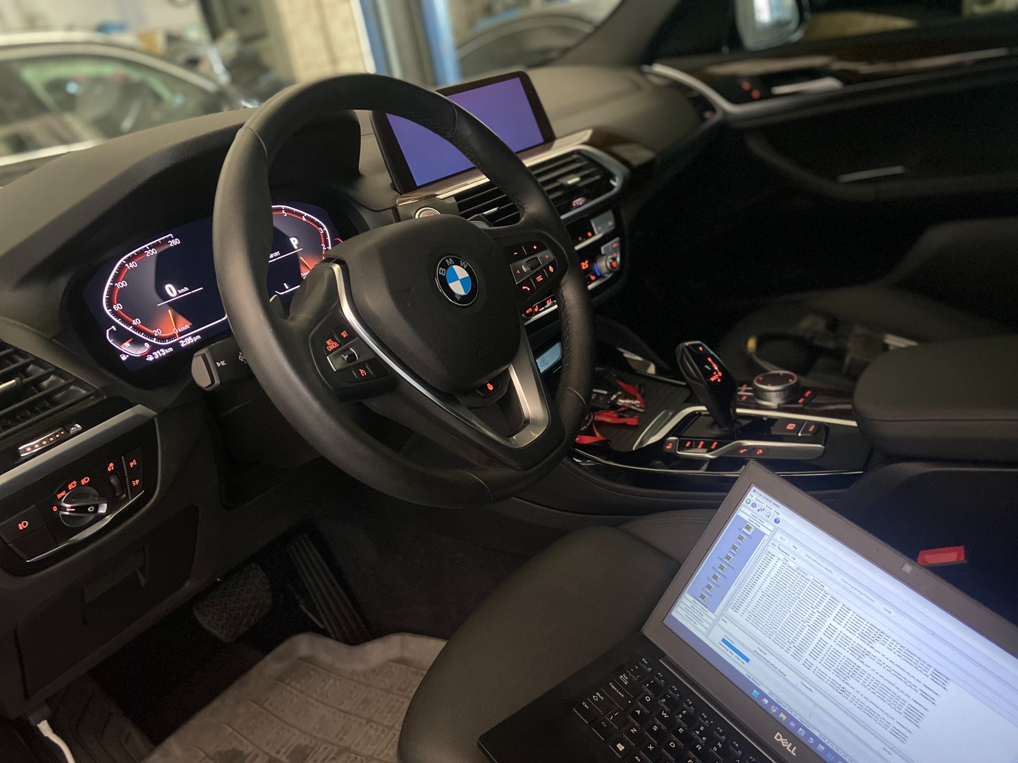 Wnętrze samochodu BMW z włączonym laptopem wyświetlającym diagnostykę pojazdu, widoczne deska rozdzielcza, kierownica z logo BMW i ekran nawigacji.