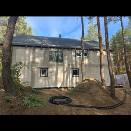 Grand Bud Masters IVAN FERENTS - Stan surowy domu z czarnymi ramami okiennymi w otoczeniu drzew w Warszawie. Widoczne czarne rury na ziemi.