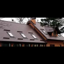 Brązowy dach z blachy z oknami dachowymi w szeregu, ceglany komin i fragment elewacji domu z oknami. Widok z dołu, pochmurne niebo w tle.