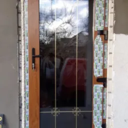 Brązowe drzwi zewnętrzne z szybą z ozdobnymi kratkami, częściowo oklejone taśmą ochronną, osadzone w murze z widoczną pianką montażową.