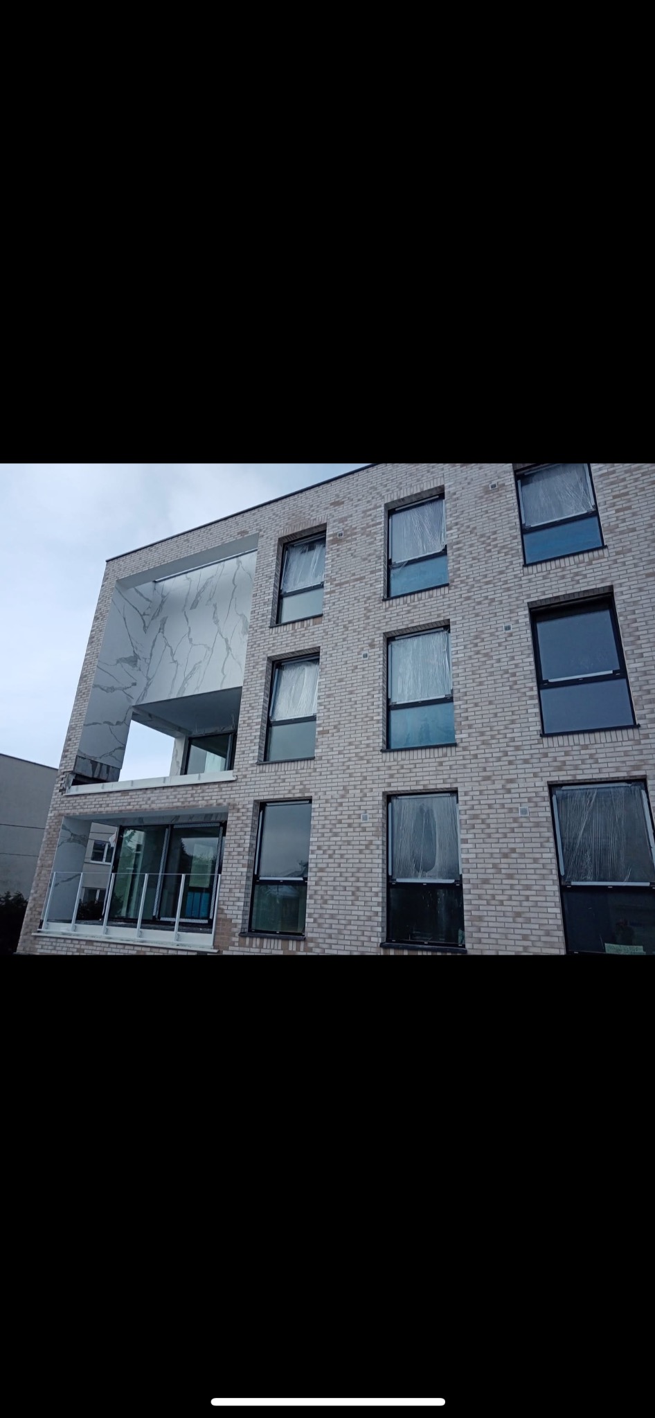 Elewacja budynku z jasnej cegły z oknami zabezpieczonymi folią i balkonem wykończonym marmurowym wzorem. Widok z dołu na nowoczesny budynek mieszkalny.