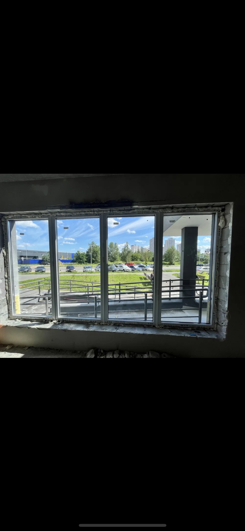 Trzyczęściowe okno PCV z widokiem na parking i zieleń, zamontowane w surowym otworze okiennym z widoczną pianką montażową i fragmentami gruzu. Dzień słoneczny.