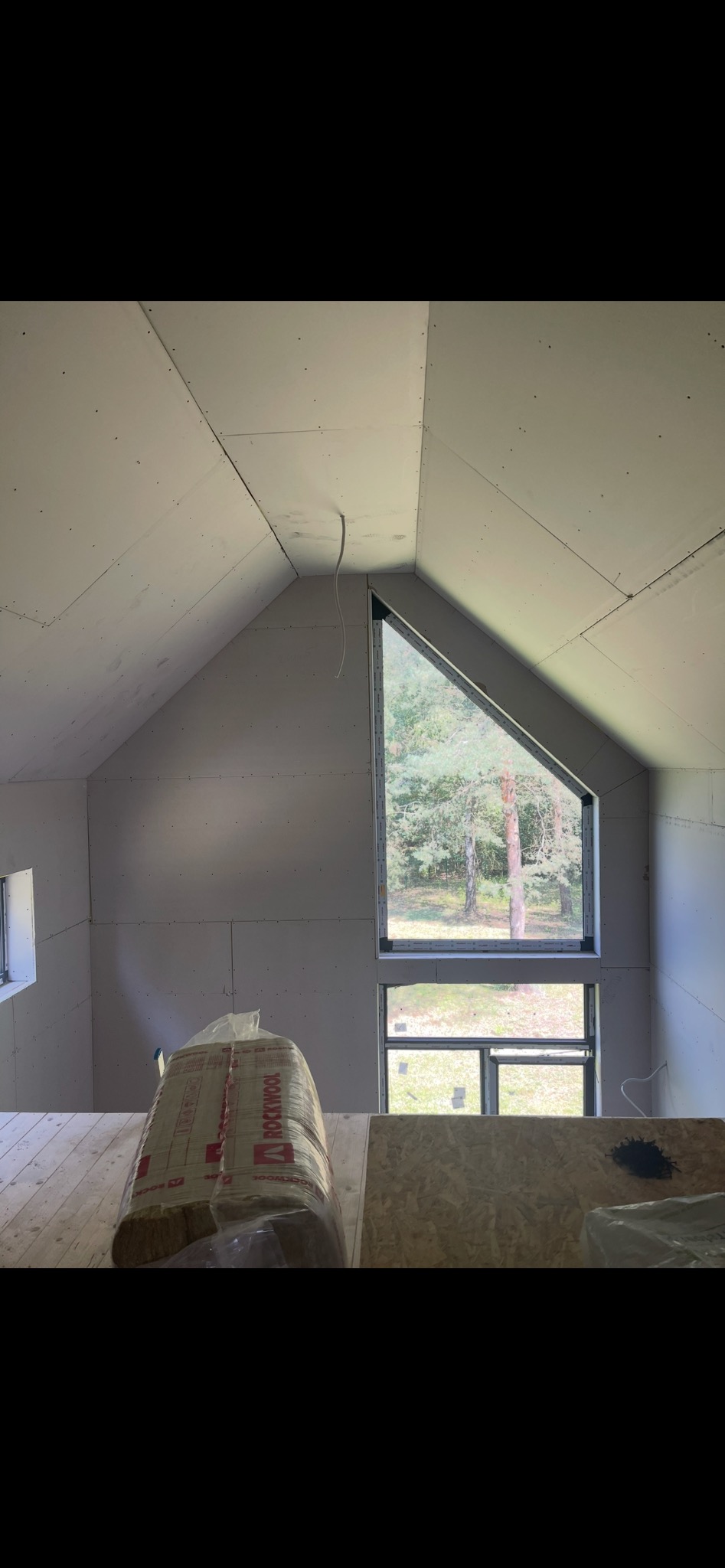 Poddasze w trakcie prac wykończeniowych: płyty gipsowe na ścianach, nietypowe okno trójkątne z widokiem na drzewa, wełna mineralna ROCKWOOL. Surowy stan przed malowaniem.