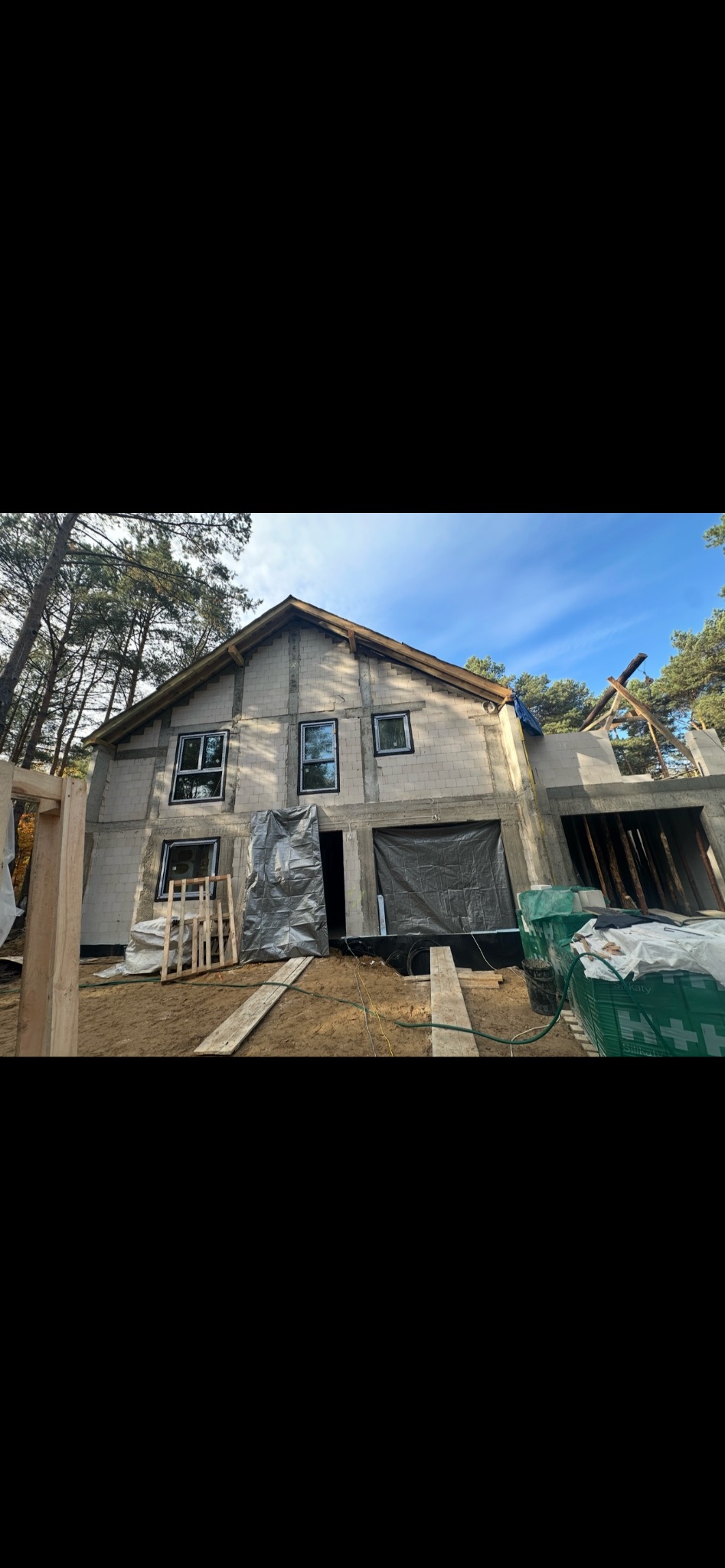 Surowy stan domu w trakcie budowy: widoczne okna PCV, ściany z bloczków, drewniana konstrukcja dachu i folia ochronna. Otoczenie leśne, jesienna aura.