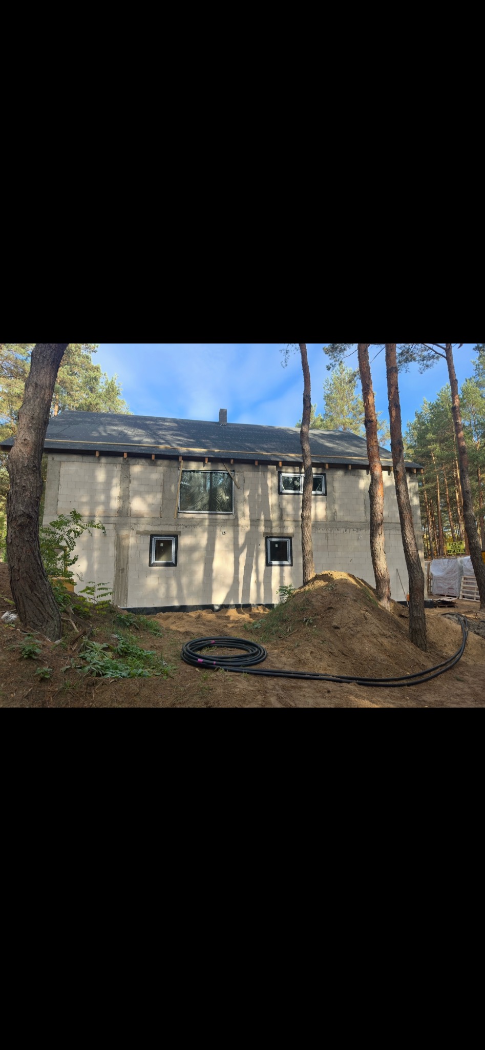 Stan surowy domu z czarnymi ramami okiennymi w otoczeniu drzew w Warszawie. Widoczne czarne rury na ziemi.