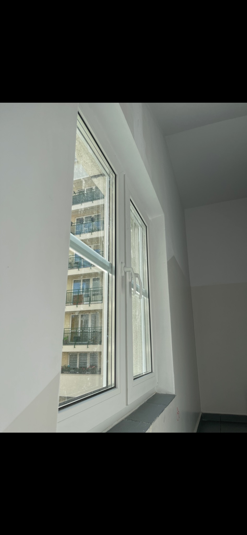 Białe okno PCV z widokiem na blok mieszkalny, uchylone skrzydło po prawej stronie. Widoczne ślady po remoncie na ścianie i parapecie.