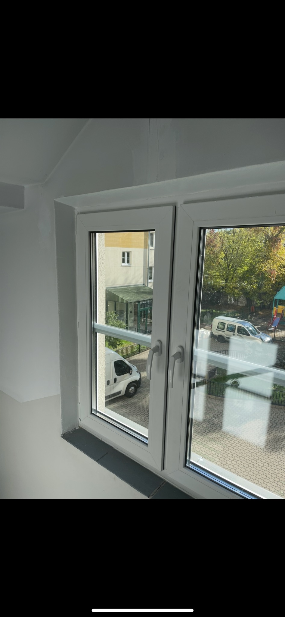 Białe okno PCV z widokiem na podwórze z zaparkowanym białym vanem. Zewnętrzna strona okna z widocznymi klamkami i szprosami.