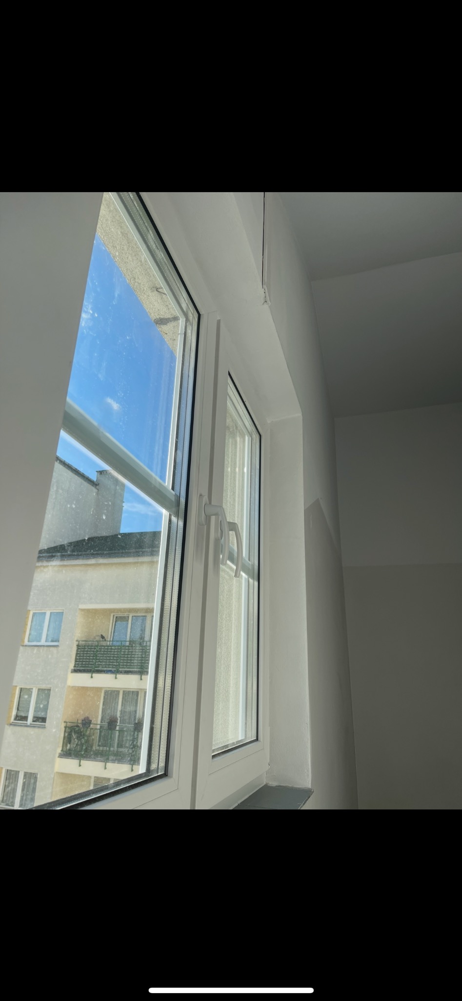Białe okno PCV z widokiem na budynek z balkonami. Okno uchylone, z widoczną klamką. Na szybie widoczne smugi. Minimalistyczne wnętrze w odcieniach bieli.