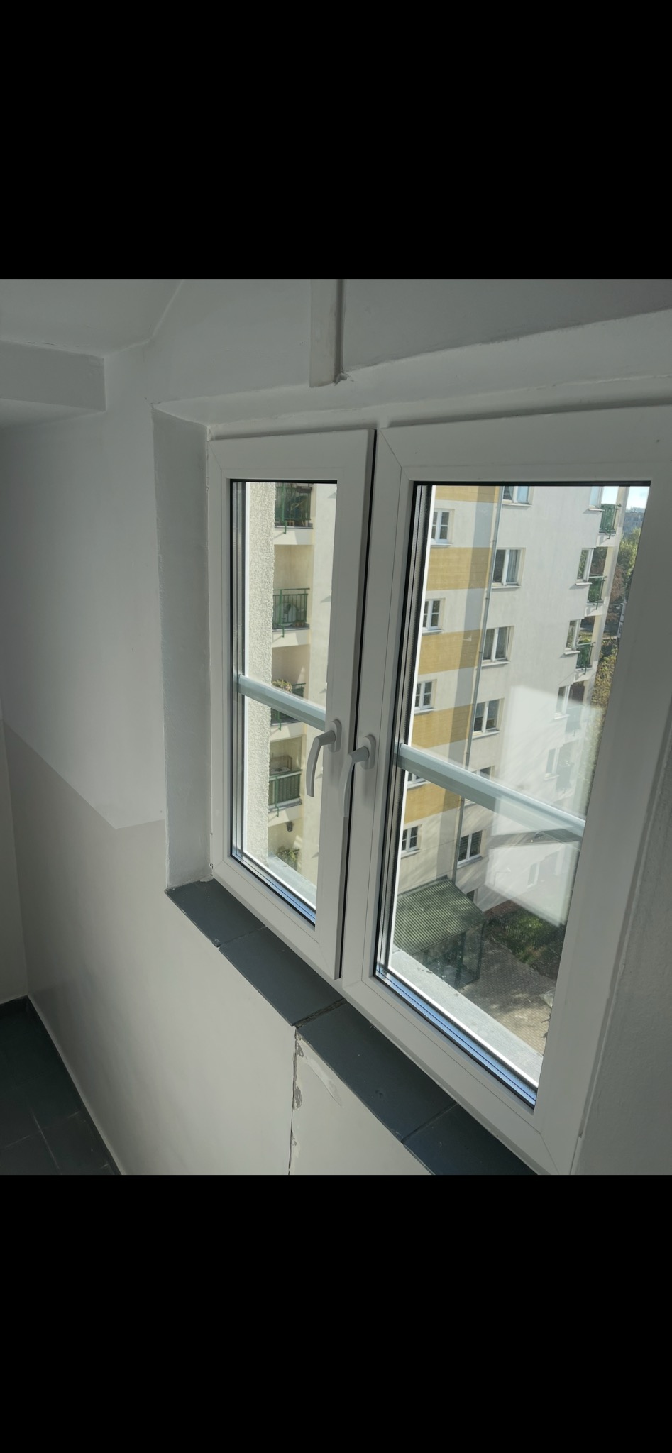 Dwuskrzydłowe okno PCV z białymi ramami, widok z klatki schodowej na budynek mieszkalny. Widoczne szprosy wewnątrz szyby okna.