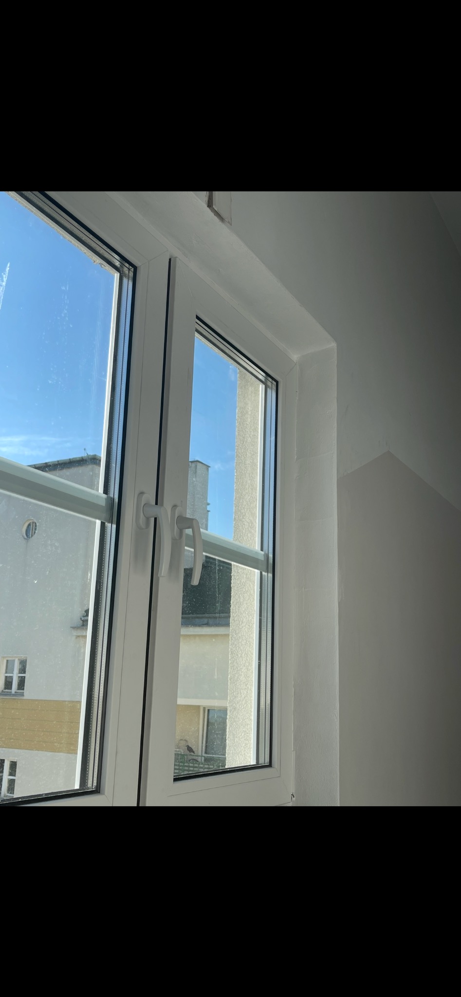 Białe, dwuskrzydłowe okno PCV z widokiem na budynek i błękitne niebo. Widoczne klamki i rama okienna. Świeżo pomalowana ściana w tle.