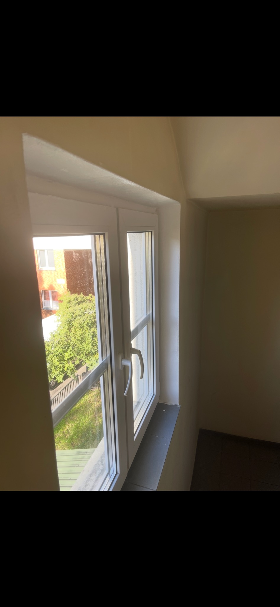 Białe okno PCV z widokiem na zieleń, uchylone, z moskitierą, w jasnym wnętrzu. Widoczny fragment parapetu i ściany.