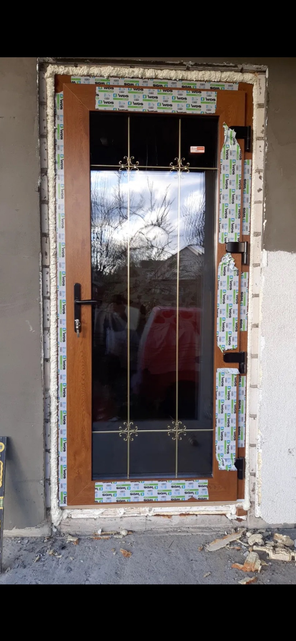 Brązowe drzwi zewnętrzne z szybą z ozdobnymi kratkami, częściowo oklejone taśmą ochronną, osadzone w murze z widoczną pianką montażową.
