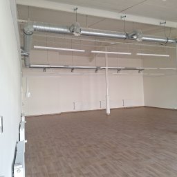 Mateusz rogala - Wnętrze pustego pomieszczenia biurowego z widocznymi elementami wentylacji pod sufitem, rzędem podłużnych lamp wiszących oraz grzejnikami na ścianach.