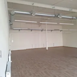 Wnętrze pustego pomieszczenia biurowego z widocznymi elementami wentylacji pod sufitem, rzędem podłużnych lamp wiszących oraz grzejnikami na ścianach.