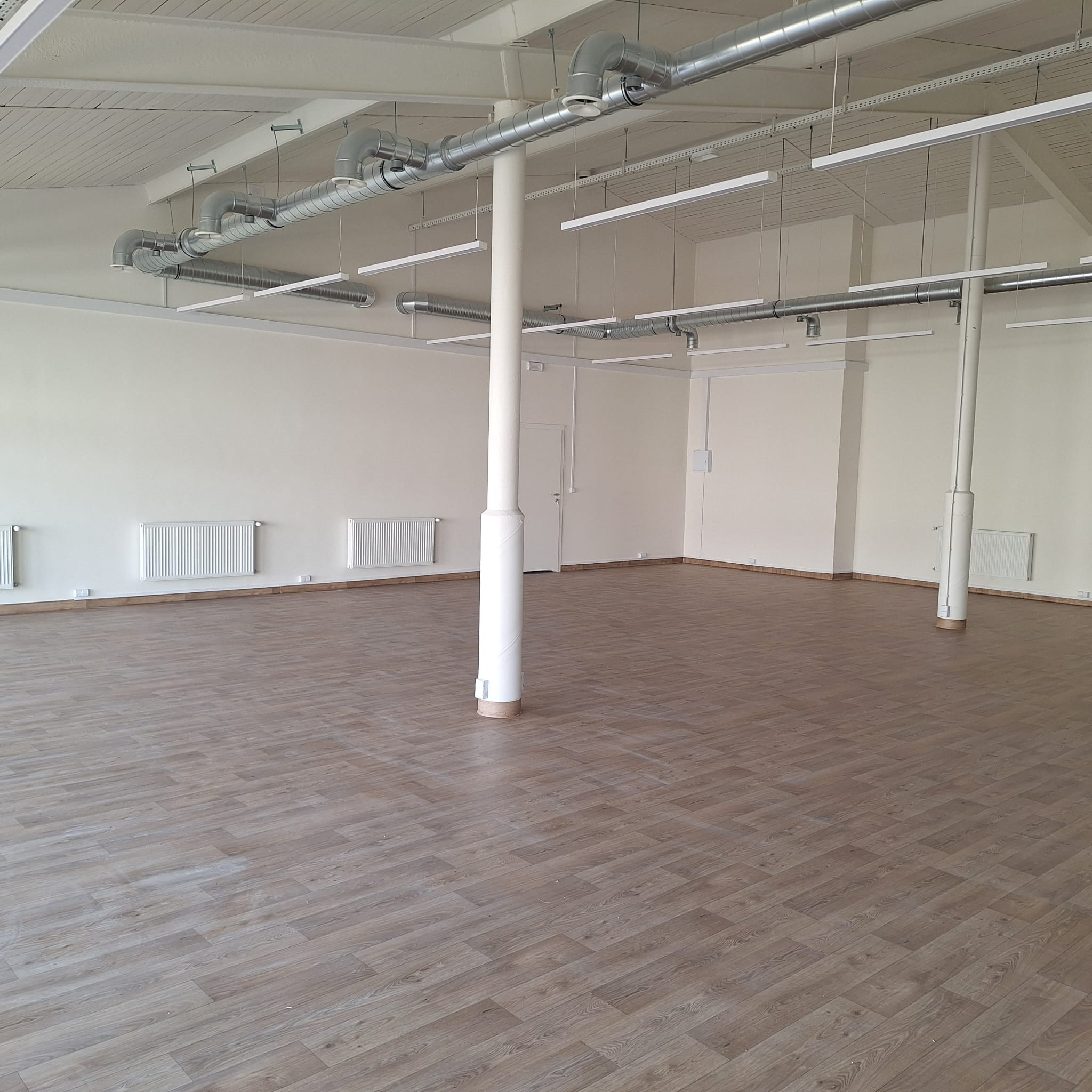 Pusta hala z drewnopodobną podłogą, białymi ścianami, grzejnikami, kolumnami i systemem wentylacji pod sufitem oraz liniowym oświetleniem.
