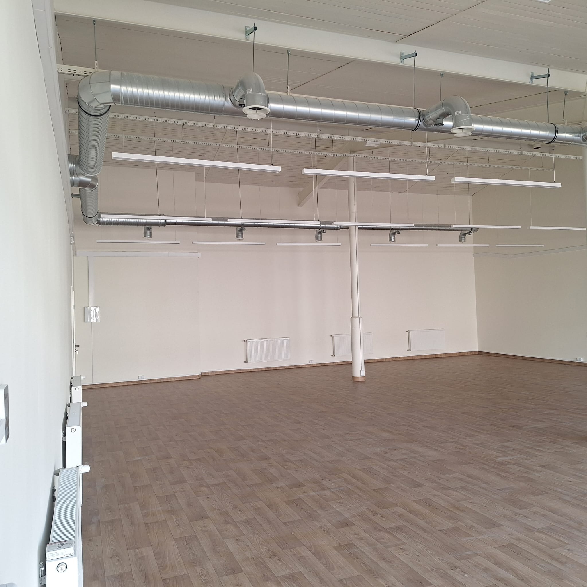 Wnętrze pustego pomieszczenia biurowego z widocznymi elementami wentylacji pod sufitem, rzędem podłużnych lamp wiszących oraz grzejnikami na ścianach.