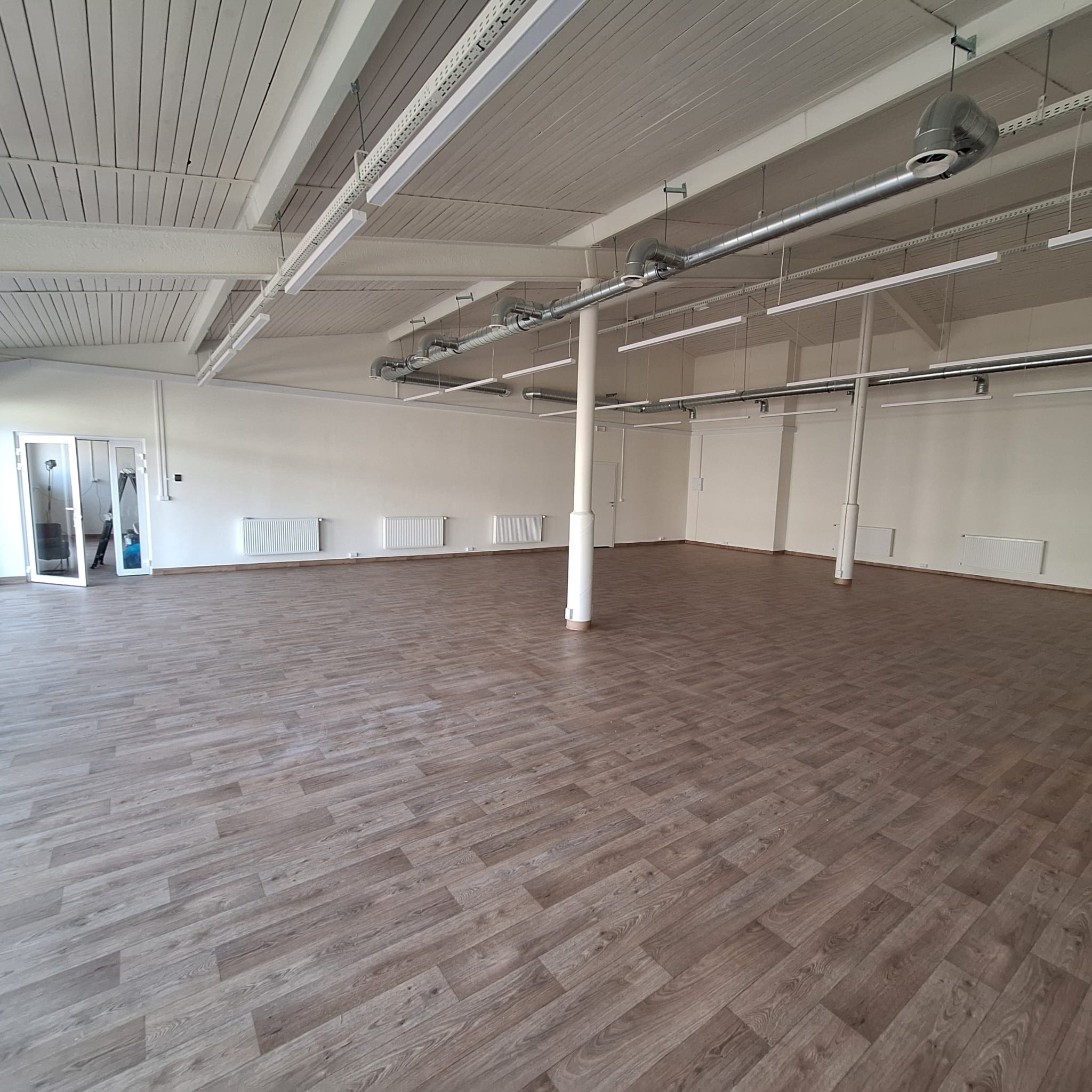 Przestronne, puste pomieszczenie z drewnianą podłogą, białymi ścianami, grzejnikami i systemem oświetlenia sufitowego z rurami wentylacyjnymi.