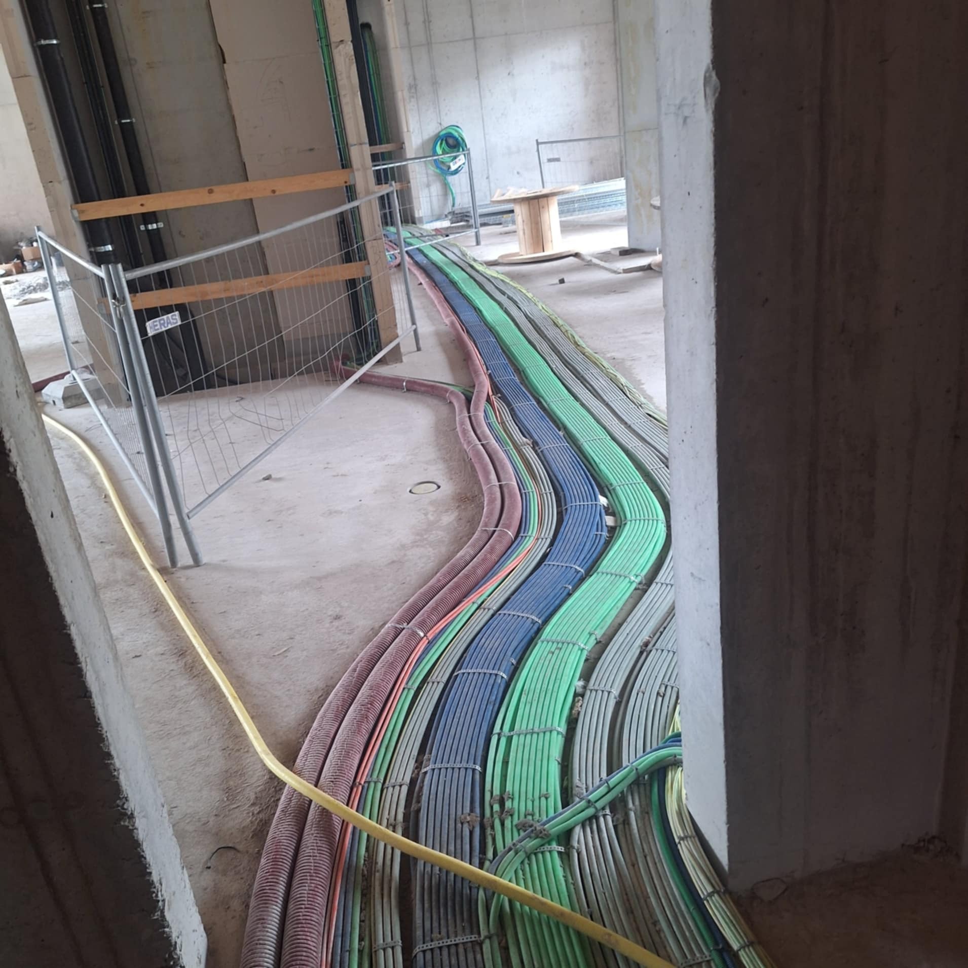 Równe rzędy kolorowych kabli elektrycznych ułożone na podłodze w surowym wnętrzu budynku w trakcie budowy, zabezpieczone metalową barierką.