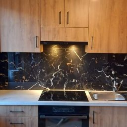 Adam Kraska Usługi remontowo-budowlane - Nowoczesna kuchnia z drewnianymi szafkami, blatem w kolorze jasnego marmuru i czarnym panelem ściennym imitującym marmur z białymi żyłami, widok na zewnątrz przez okno.