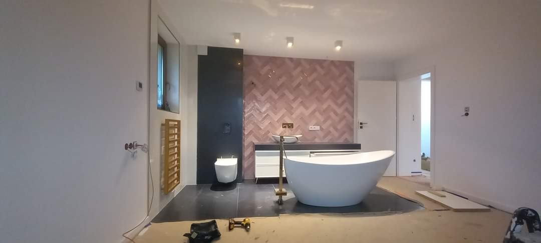 Nowoczesna łazienka w trakcie remontu: różowa mozaika w jodełkę, wolnostojąca wanna, czarne płytki podłogowe, grzejnik drabinkowy. Widoczne narzędzia i niedokończone wykończenia.