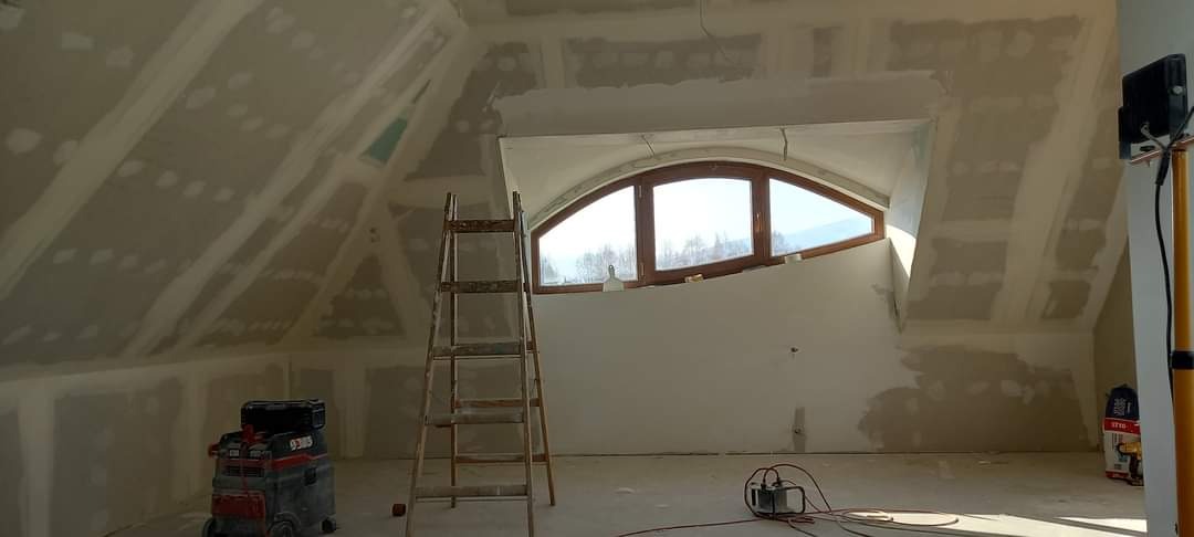 Poddasze w trakcie wykańczania: widoczne płyty kartonowo-gipsowe na skosach, częściowo zaszpachlowane, drewniana drabina oparta o ścianę, okno w kształcie łuku, odkurzacz przemysłowy i lampa...