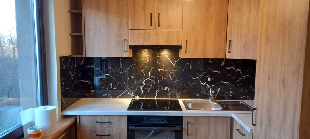 Nowoczesna kuchnia z drewnianymi szafkami, blatem w kolorze jasnego marmuru i czarnym panelem ściennym imitującym marmur z białymi żyłami, widok na zewnątrz przez okno.