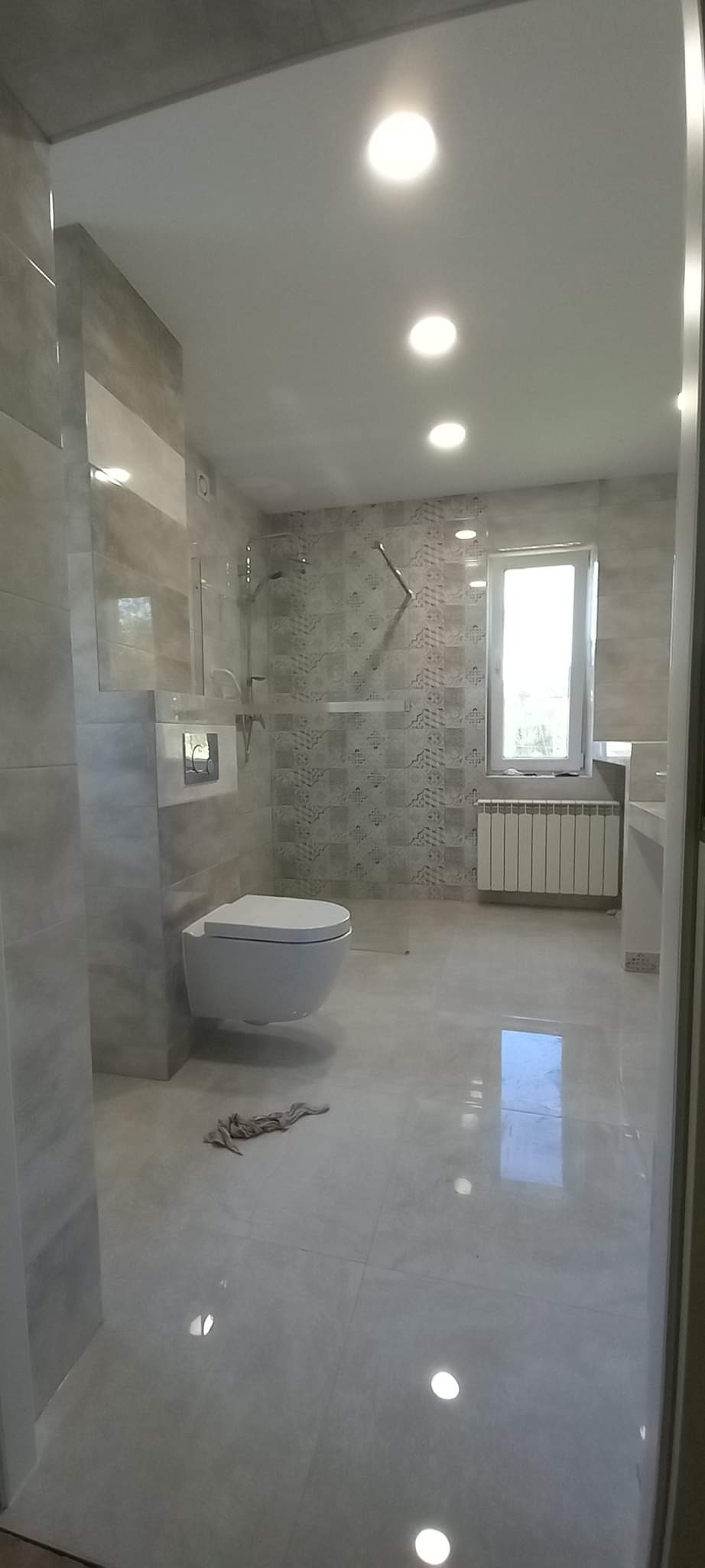 Nowoczesna łazienka z podwieszaną toaletą, kabiną prysznicową typu walk-in i jasnymi płytkami na ścianach i podłodze. Na podłodze leży szmatka. Widoczne okno z grzejnikiem pod spodem.