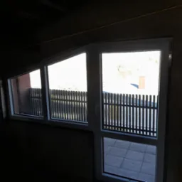 Białe okna zamontowane w surowym wnętrzu, widok na zewnętrzny taras z drewnianą balustradą. Nierówne wykończenie ścian kontrastuje z nowymi oknami.