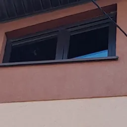 Szare okno na elewacji dwukolorowego budynku. Widoczne odbicie światła w szybie i fragment parapetu. Ujęcie z dołu, blisko okna.