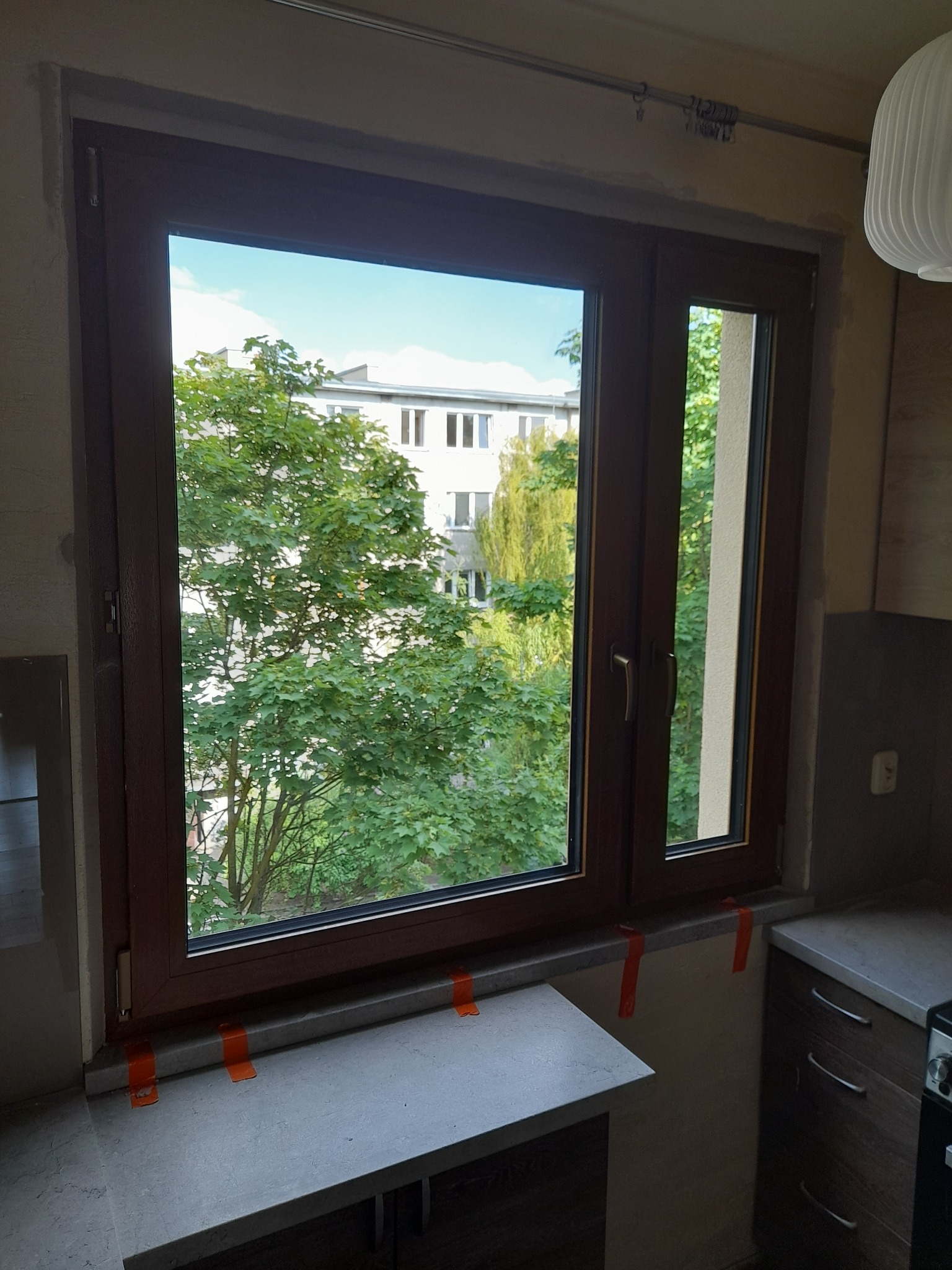 Brązowe okno z widokiem na zieleń i budynek, zabezpieczone pomarańczową taśmą. Widoczne fragmenty szafek kuchennych i lampy. Okno dwuskrzydłowe, z jednym wąskim skrzydłem.