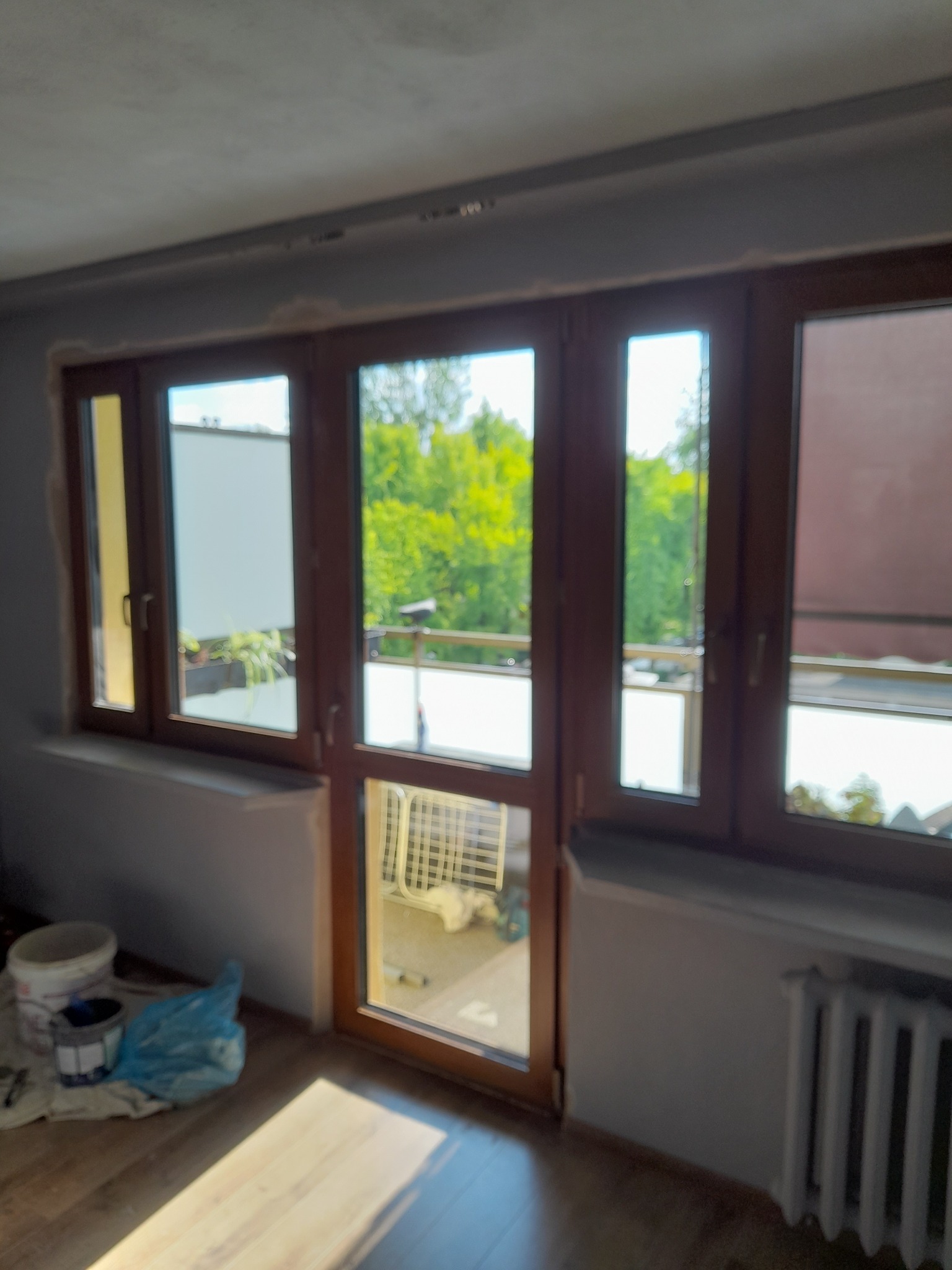 Montaż okien balkonowych z brązowymi ramami; widok na zieleń za oknem. Na podłodze widoczne narzędzia i farby. Grzejnik pod oknem.