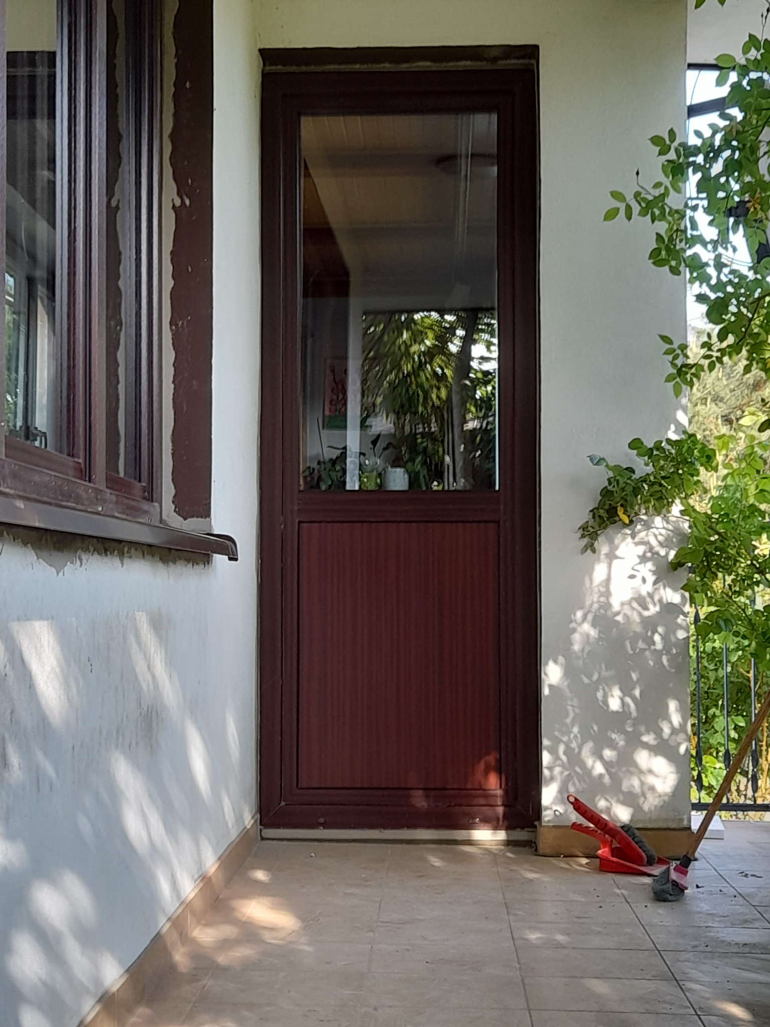 Brązowe okno PCV z szybą, widoczne z zewnątrz, z elementami roślinności po prawej stronie i śladami użytkowania na ścianie.