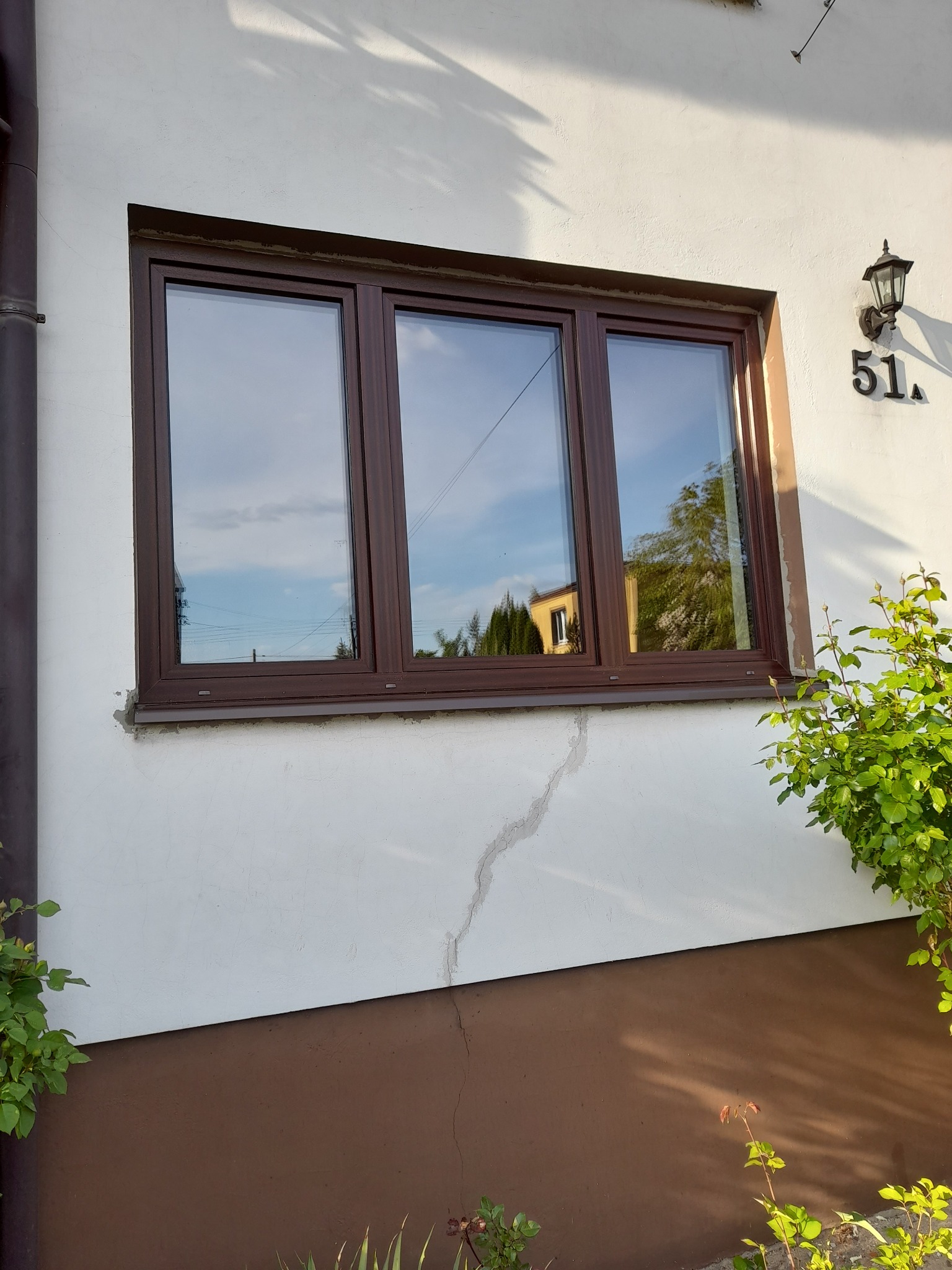 Brązowe okno PCV z trzema szybami, zamontowane w białym budynku z numerem 51. Widoczna elewacja z pęknięciem, lampa zewnętrzna i zieleń. W szybach odbija się niebo.