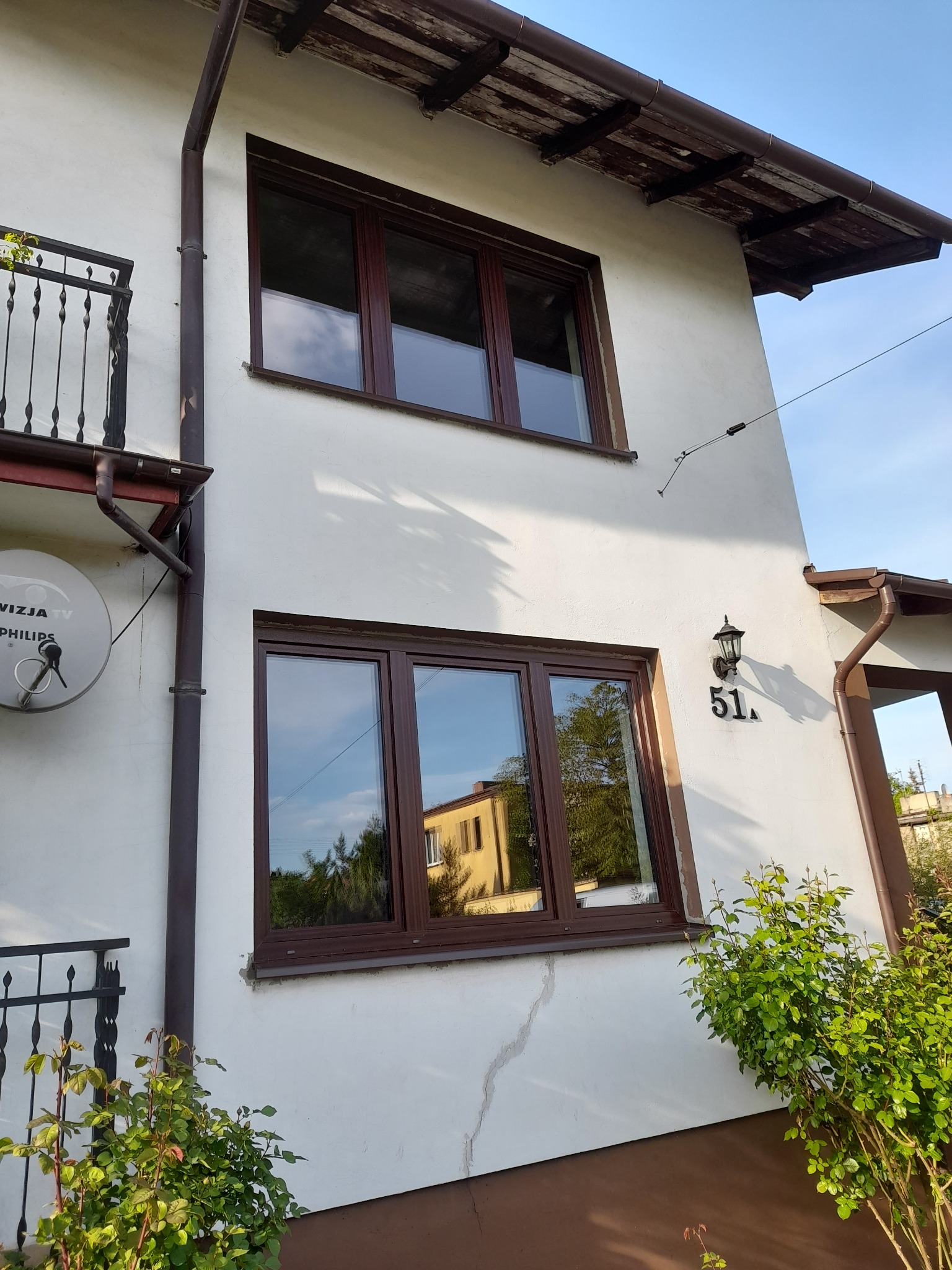 Brązowe okna w białym domu z numerem 51, z widocznym odbiciem budynku w szybach, balkonem i fragmentem rynny. Ujęcie z dołu, w słoneczny dzień.