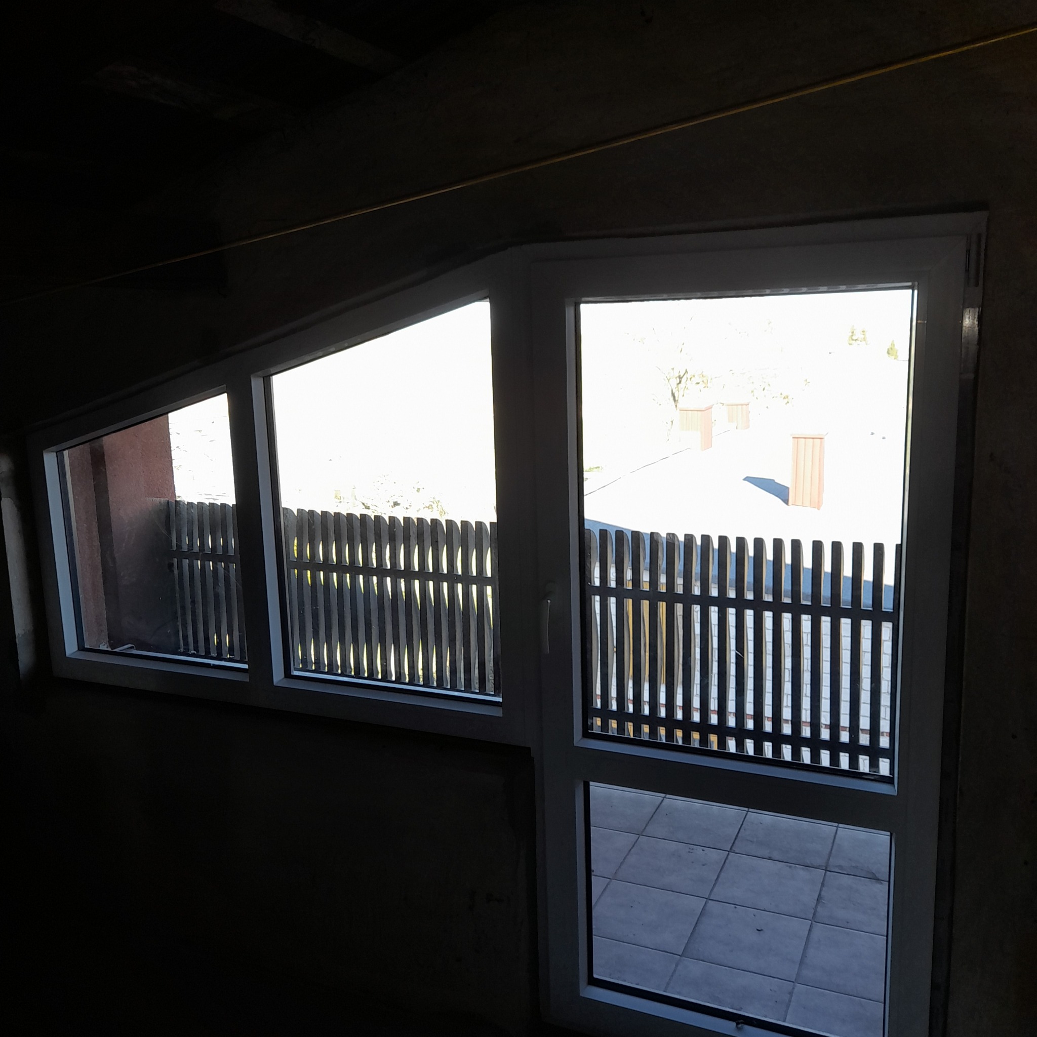 Białe okna zamontowane w surowym wnętrzu, widok na zewnętrzny taras z drewnianą balustradą. Nierówne wykończenie ścian kontrastuje z nowymi oknami.