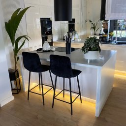 Starz-Drew - Wnętrze nowoczesnej, jasnej kuchni z wyspą, czarnymi hokerami i podświetleniem LED pod szafkami, widok na sprzęt AGD w zabudowie.