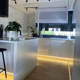 Starz-Drew - Nowoczesna, jasna kuchnia z wyspą wykończoną marmurem, oświetleniem LED pod szafkami i roślinami doniczkowymi na blacie, widok na okno z roletami i firanką.