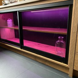 Starz-Drew - Szafka z przesuwnymi drzwiami, wykonana z drewna w ciepłym odcieniu, oświetlona fioletowym światłem, z dekoracyjnymi przedmiotami na półkach.